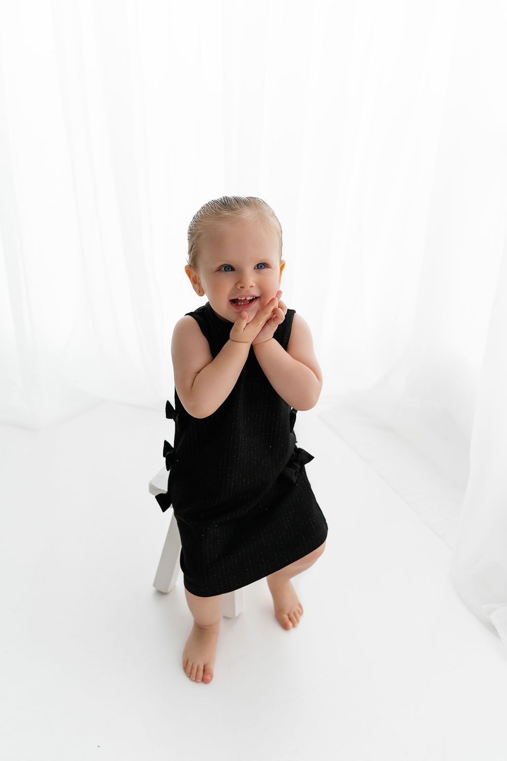 Sweet toddler in black sleeveless dress standing barefoot and giggling against white backdrop.