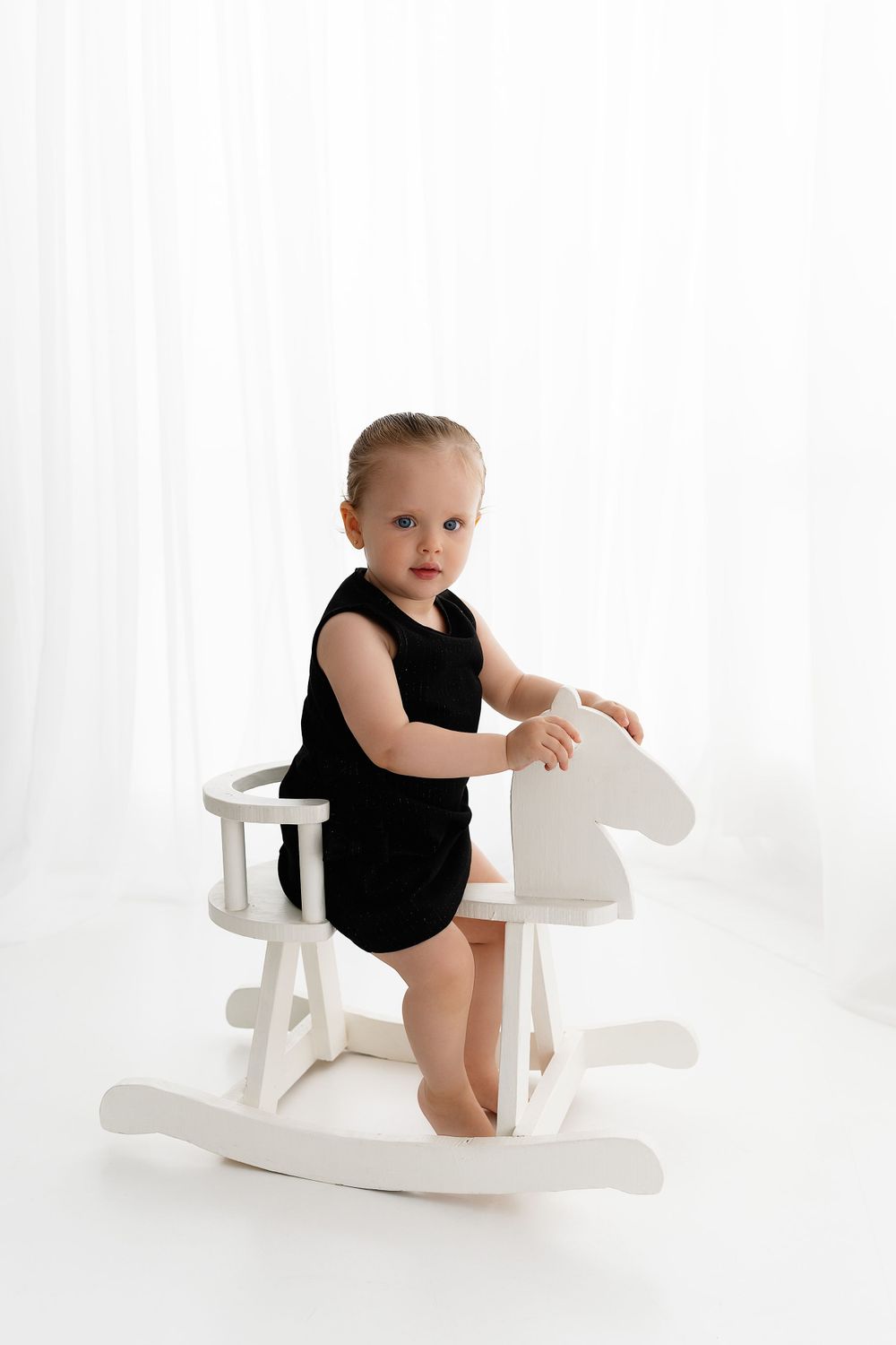 Small child wearing black leotard stands on white rocking horse toy against white backdrop in studio setting.