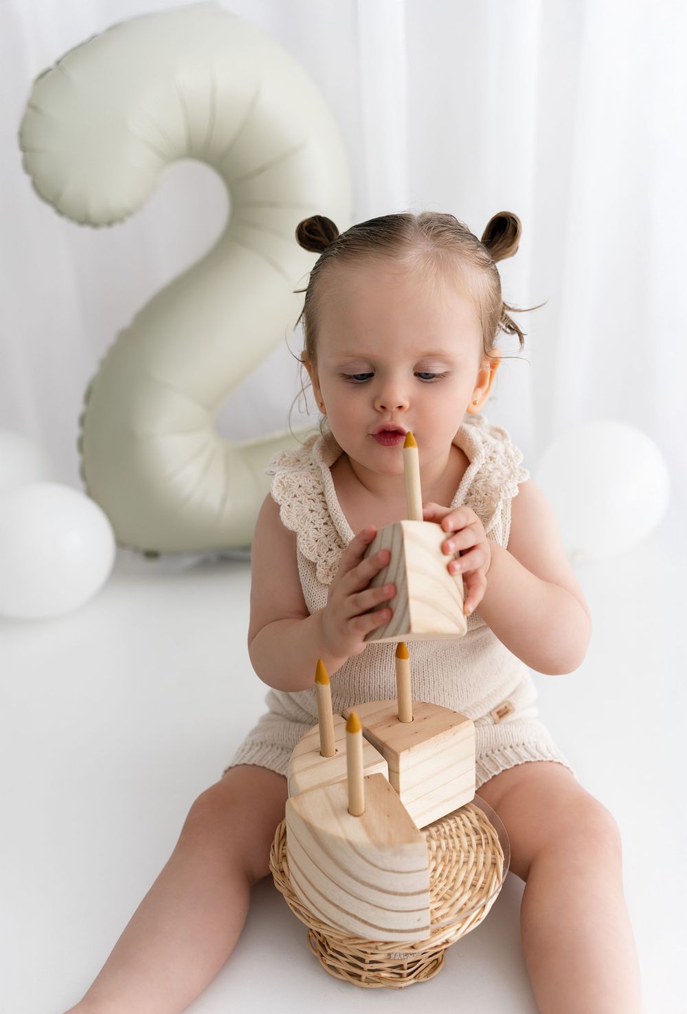 Sweet toddler in lace romper drinks from bottle while sitting beside large white number 2 birthday decoration.