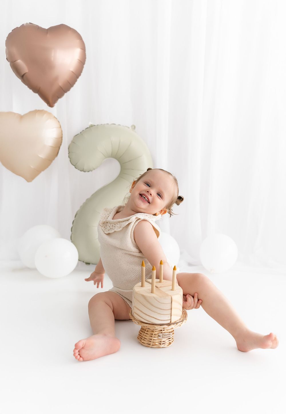 Second birthday celebration setup with rose gold heart balloons, sage number 2 balloon and minimal cake decoration on white.