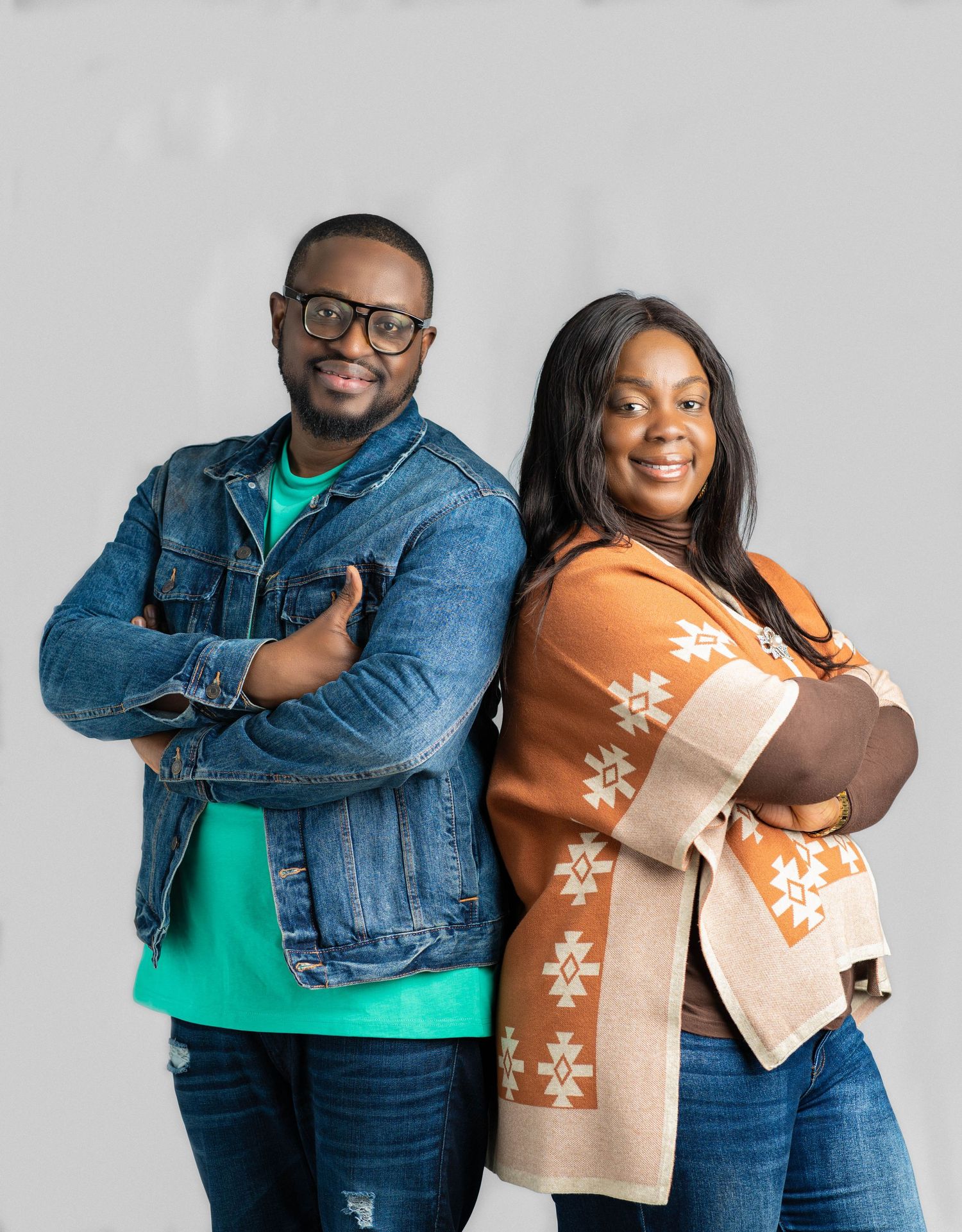 Two people stand close together against a light background, wearing denim and a patterned cardigan.