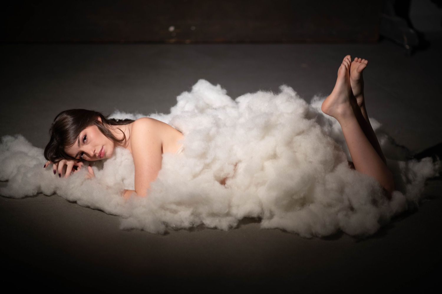 Woman reclining gracefully on a pile of soft white fluffy material in a dark studio.