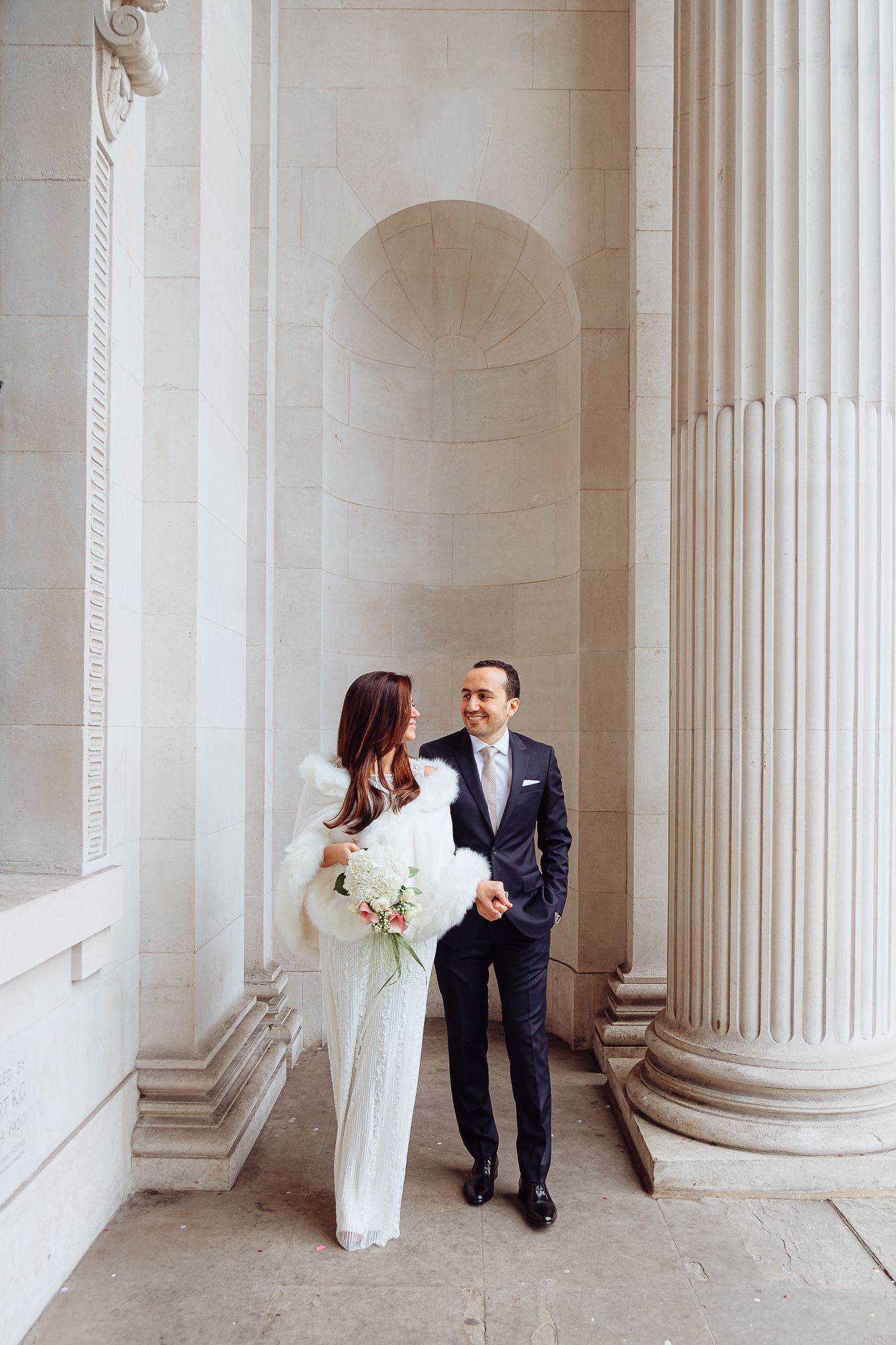 A romantic wedding couple stands between elegant white stone columns in a grand classical building.