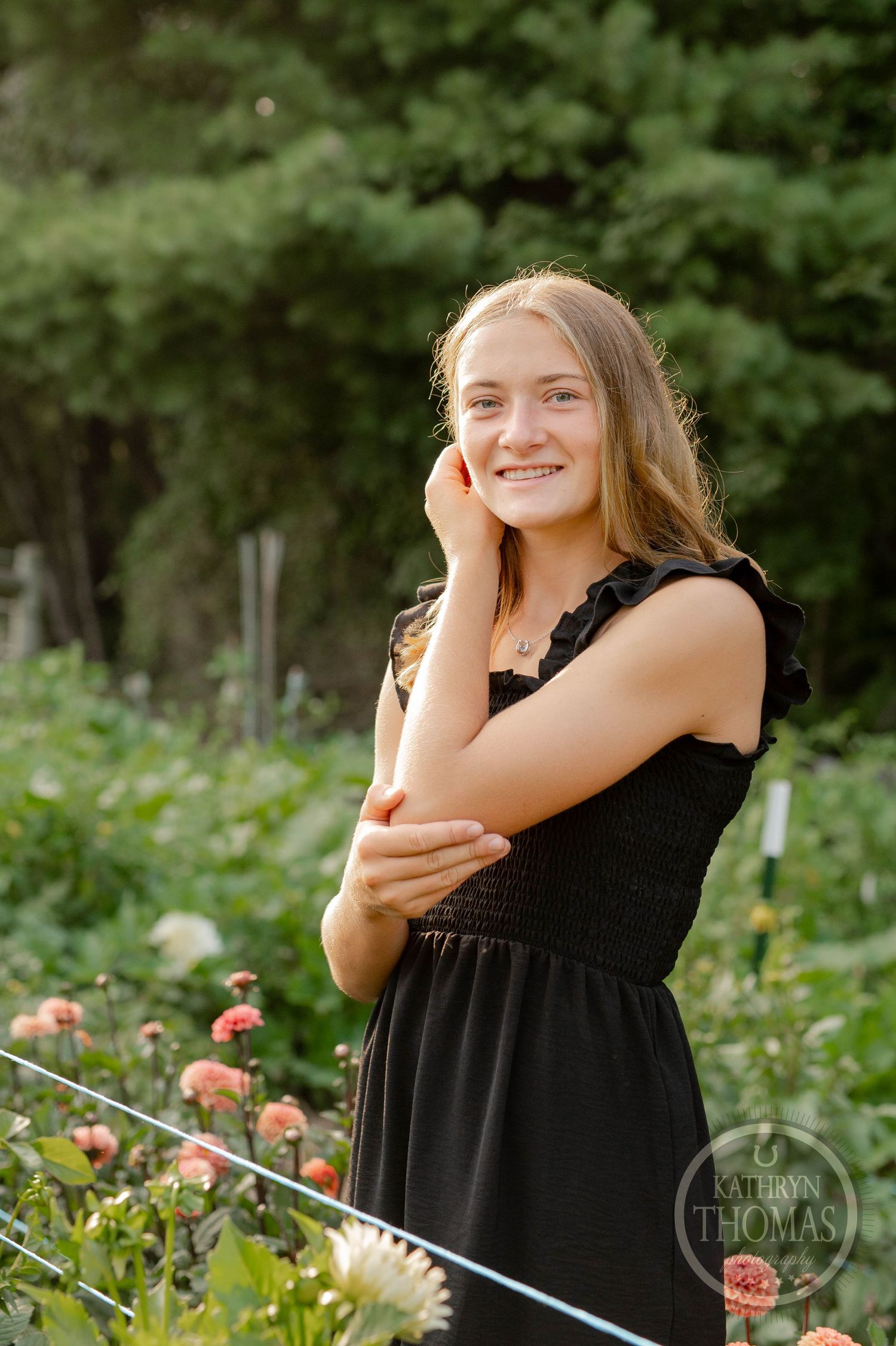 Young woman in black dress smiles in a garden filled with coral-colored flowers and greenery.