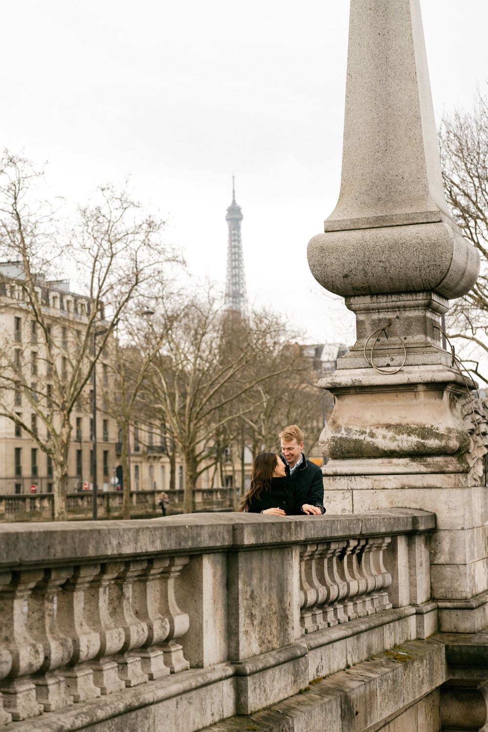 Person in black coat stands on ornate stone bridge with iconic Eiffel Tower visible in misty Paris background.