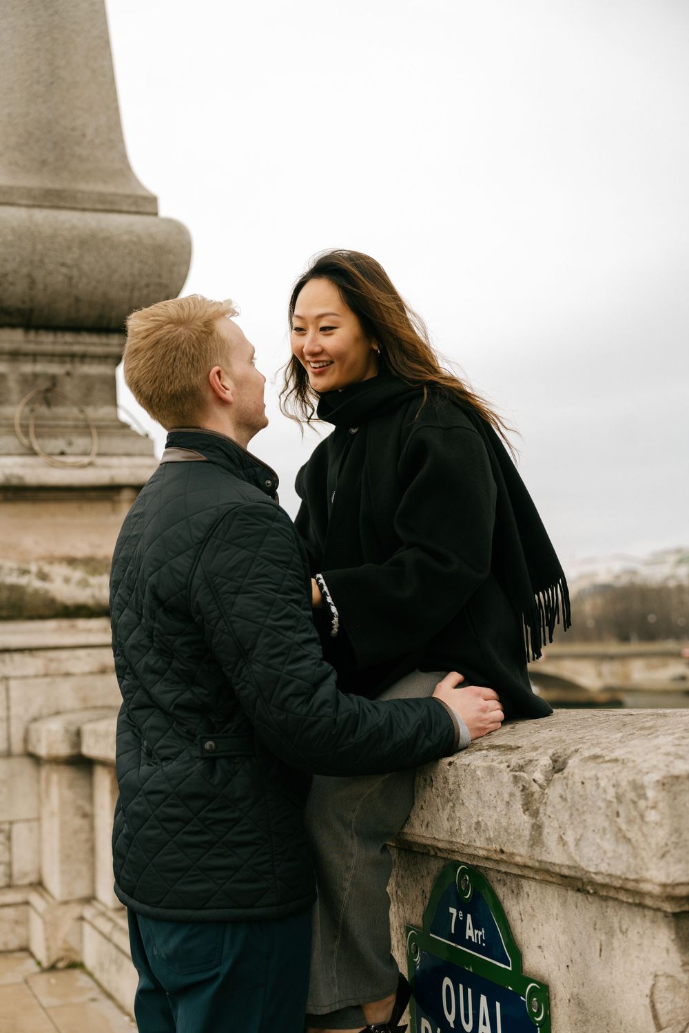 A couple shares a romantic moment together on a stone balcony overlooking a cloudy cityscape.