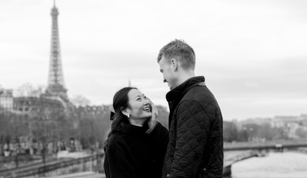 A romantic couple shares a tender moment in front of the Eiffel Tower in black and white Paris cityscape.