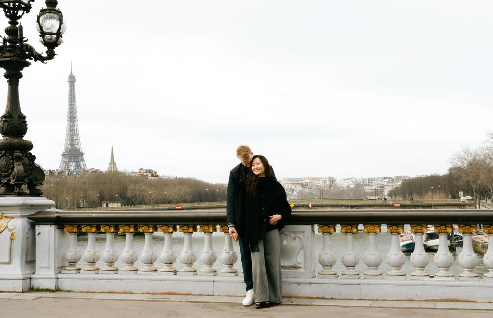 Couple standing on Pont Alexandre III bridge in Paris with the Eiffel Tower visible in the distance.