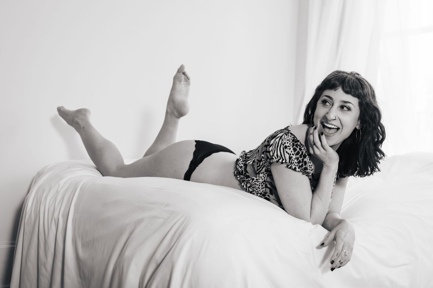 Black and white boudoir photo showing a playful pose on a bed with a leopard print top and dark leggings.