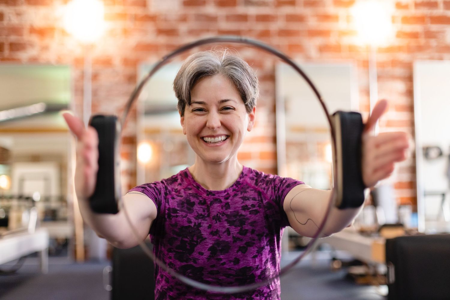 Someone wearing a purple tank top performs arm exercises at a fitness center with exposed brick walls and warm lighting.
