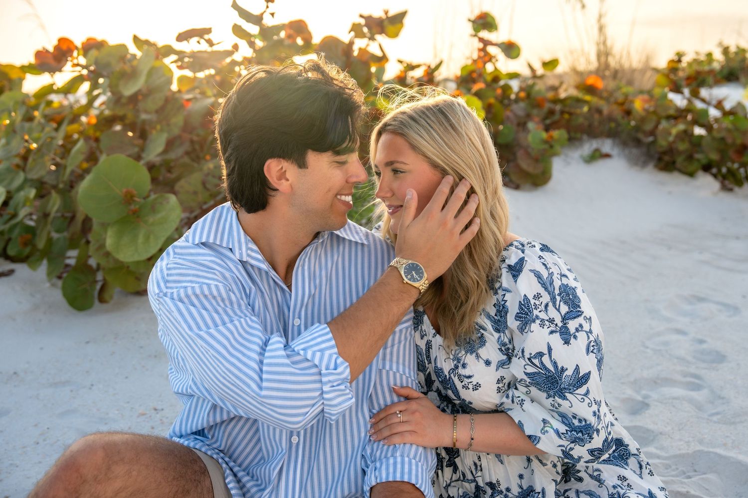 Romantic couple sitting on white sand beach at sunset, man gently cupping womans face.
