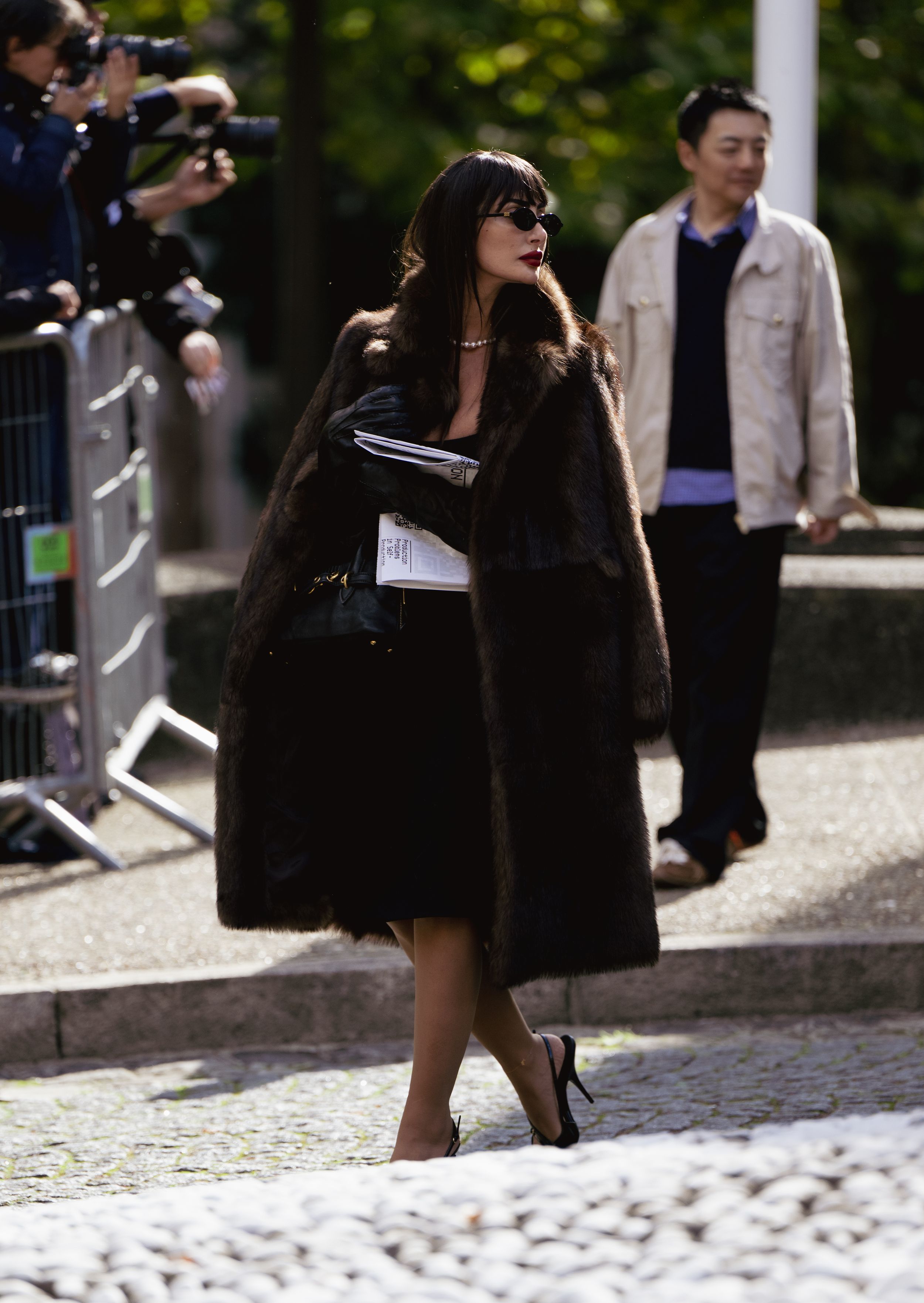 Stylish woman in black fur coat and sunglasses walking past photographers at the Miu Miu SS25 Show