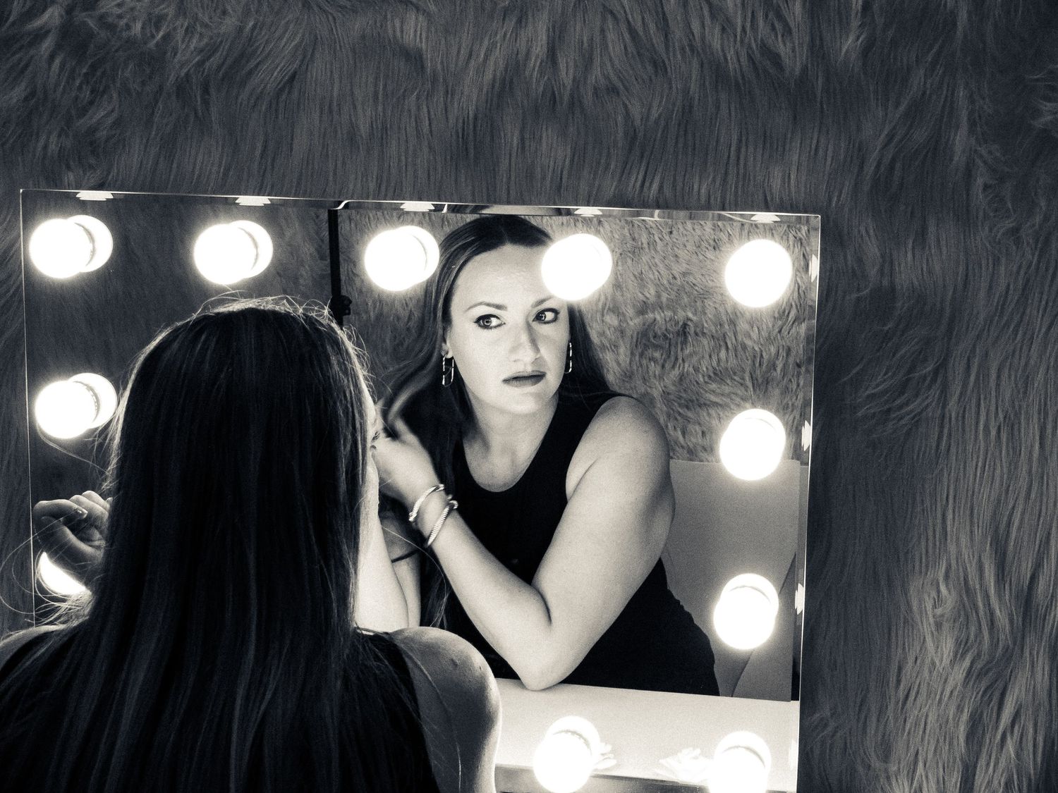 Black and white photo of a dressing room mirror with bright vanity lights reflecting a figure in dark clothing.