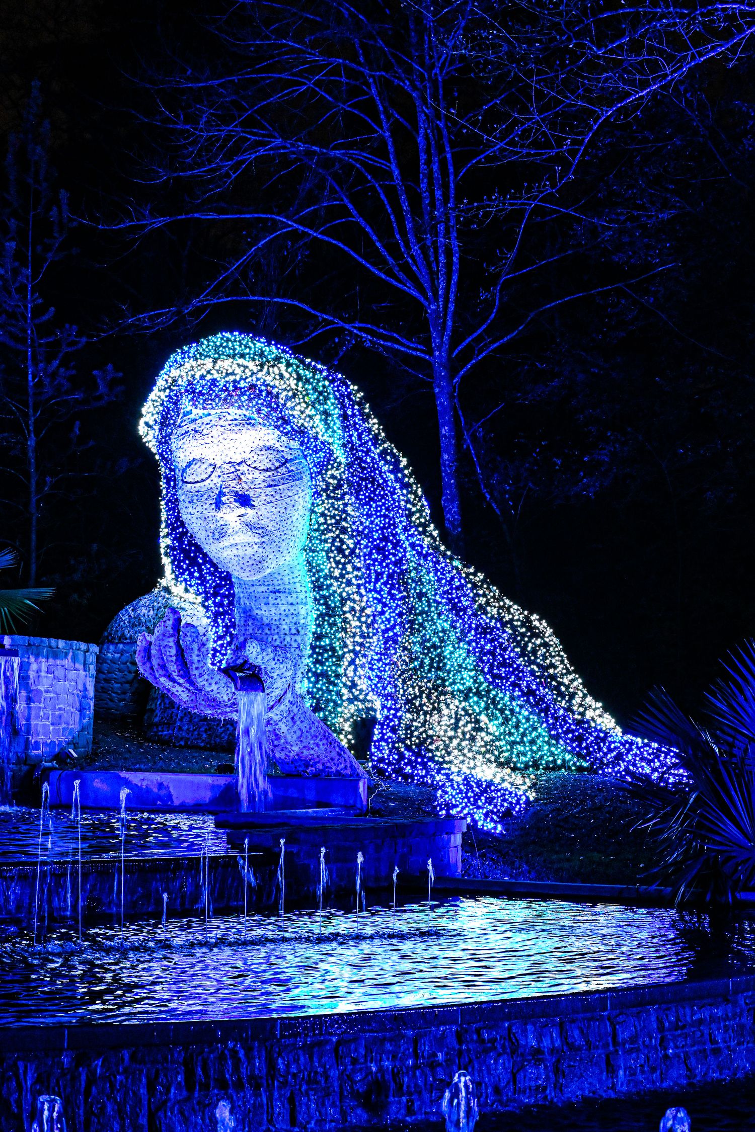 A luminous blue light sculpture in the shape of a figure head emerges from a dark garden with reflections on water below.