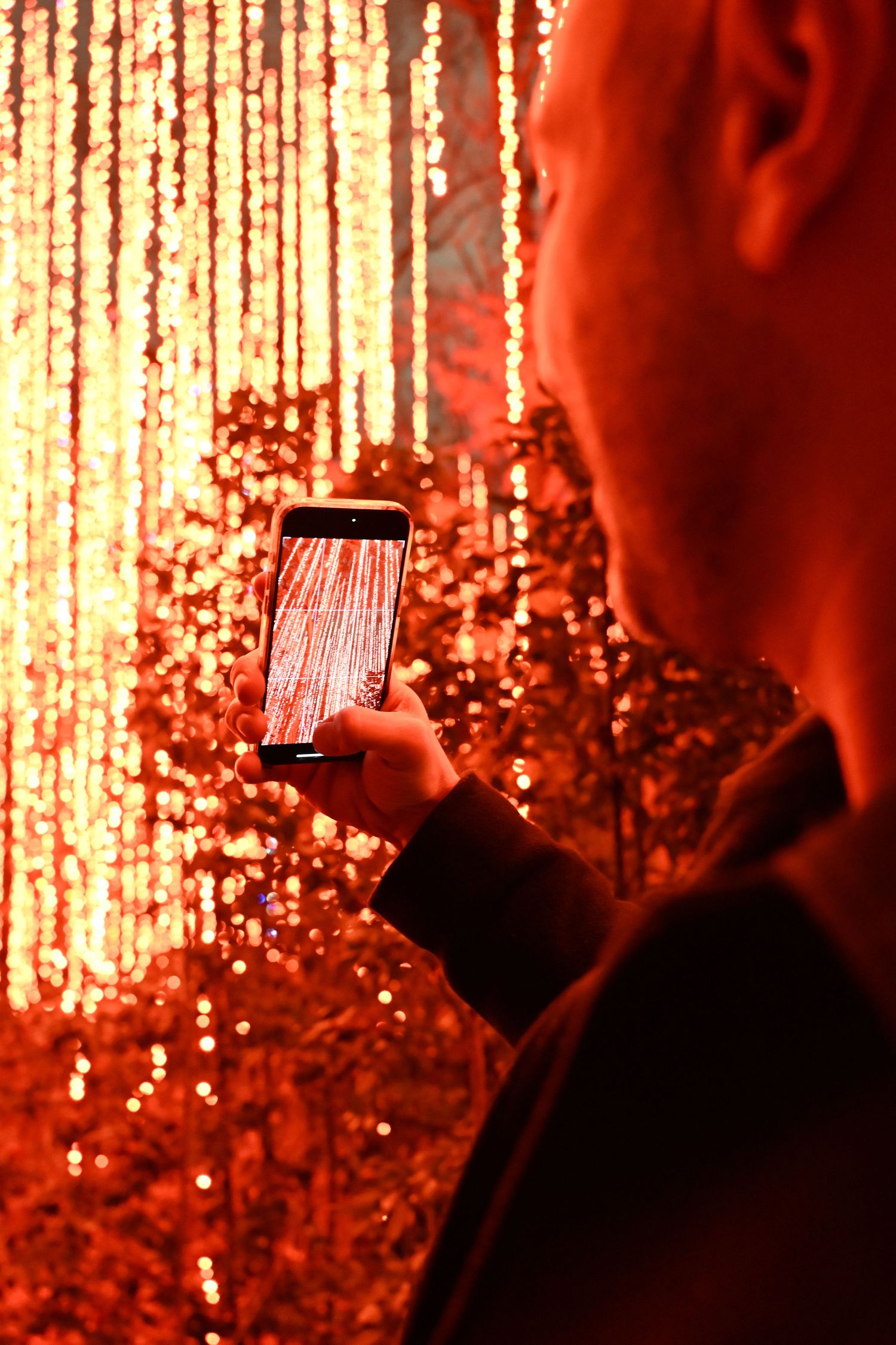 Person taking photo of glowing red string lights installation with smartphone in dark setting.