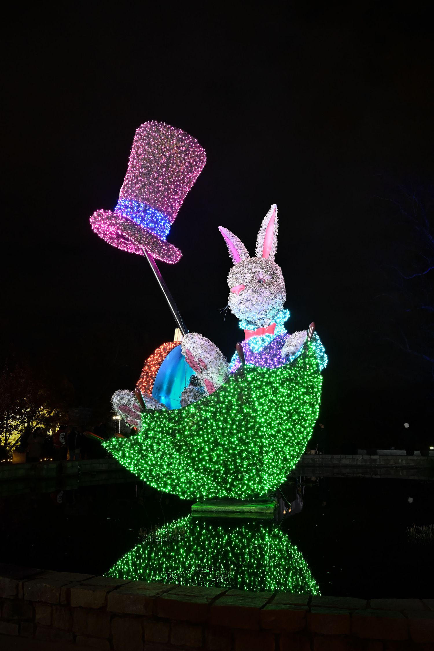 Illuminated holiday display featuring a bunny in a green sleigh with a glowing pink top hat and blue accents at night.