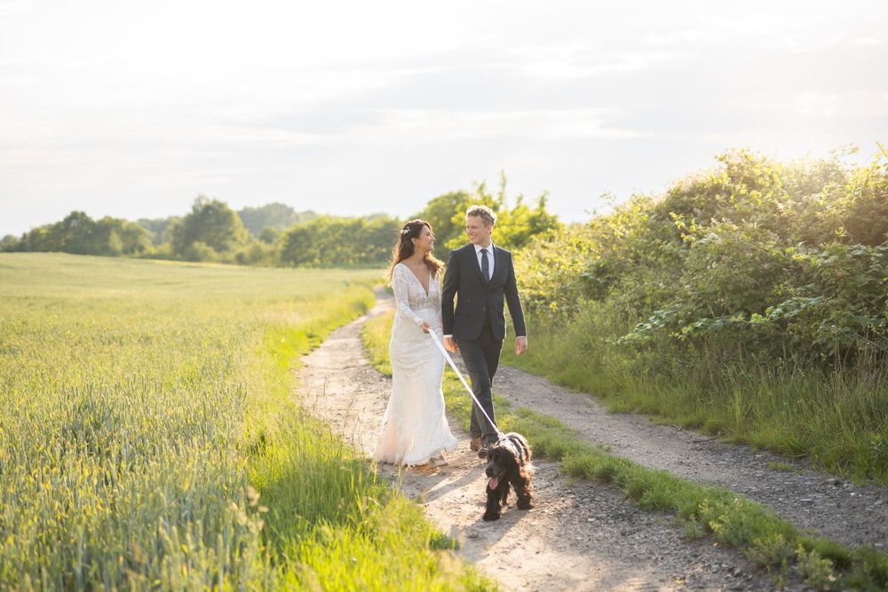Couple walking with dog on countryside path during golden hour wedding photoshoot.