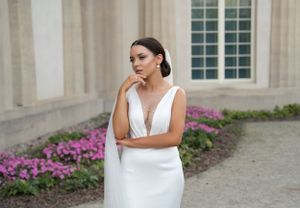 Elegant bride in white v-neck dress posing against stone building with purple flowers.
