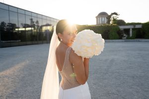 Backlit bride holding white peony bouquet outside modern building at sunset.