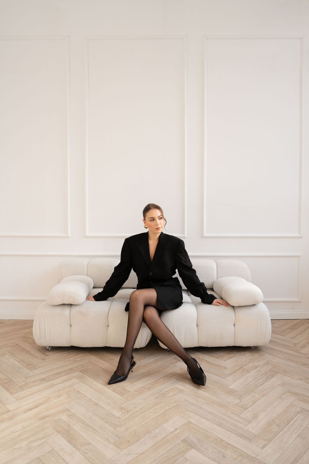 Chic black business attire styled with stockings on white modern sofa against paneled wall.