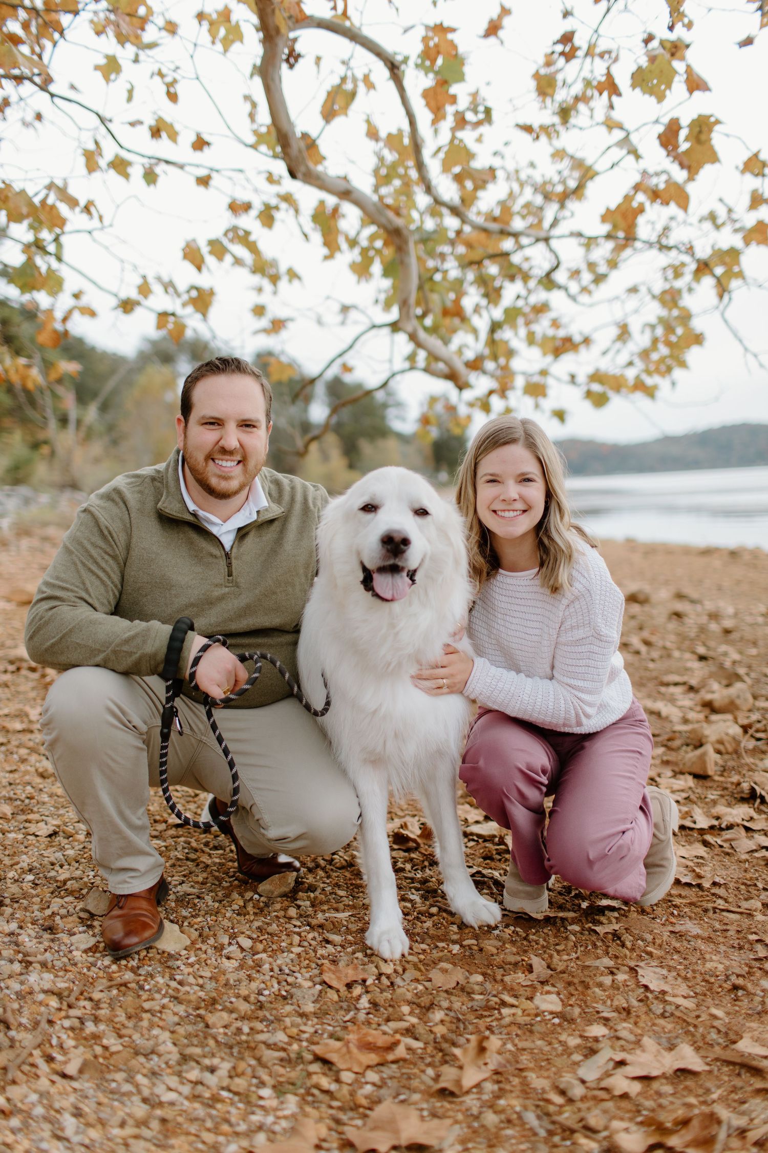 A happy white fluffy dog poses with people on fallen autumn leaves near a lake.