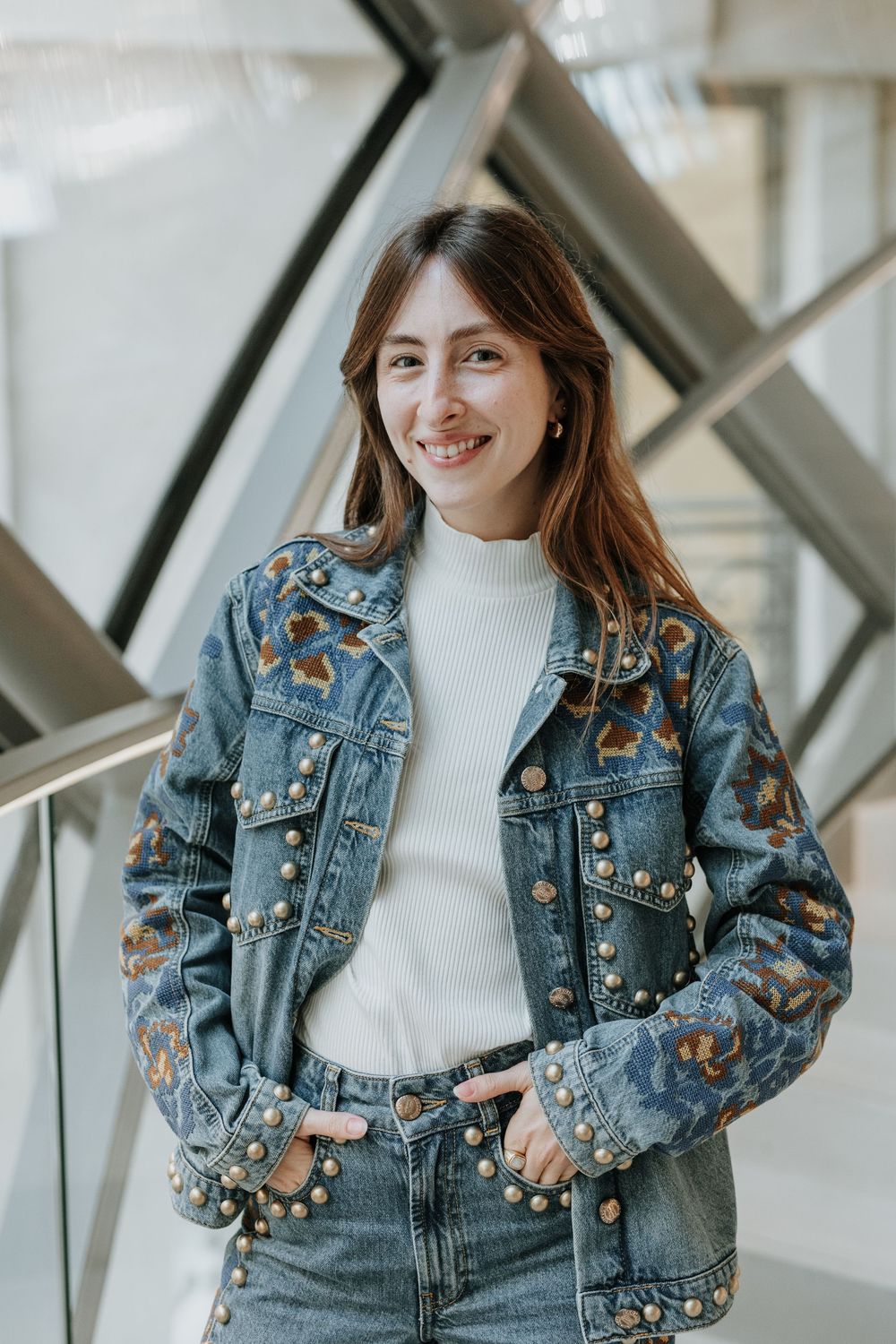 Person wearing embroidered butterfly denim jacket with white turtleneck sweater poses in bright indoor setting.