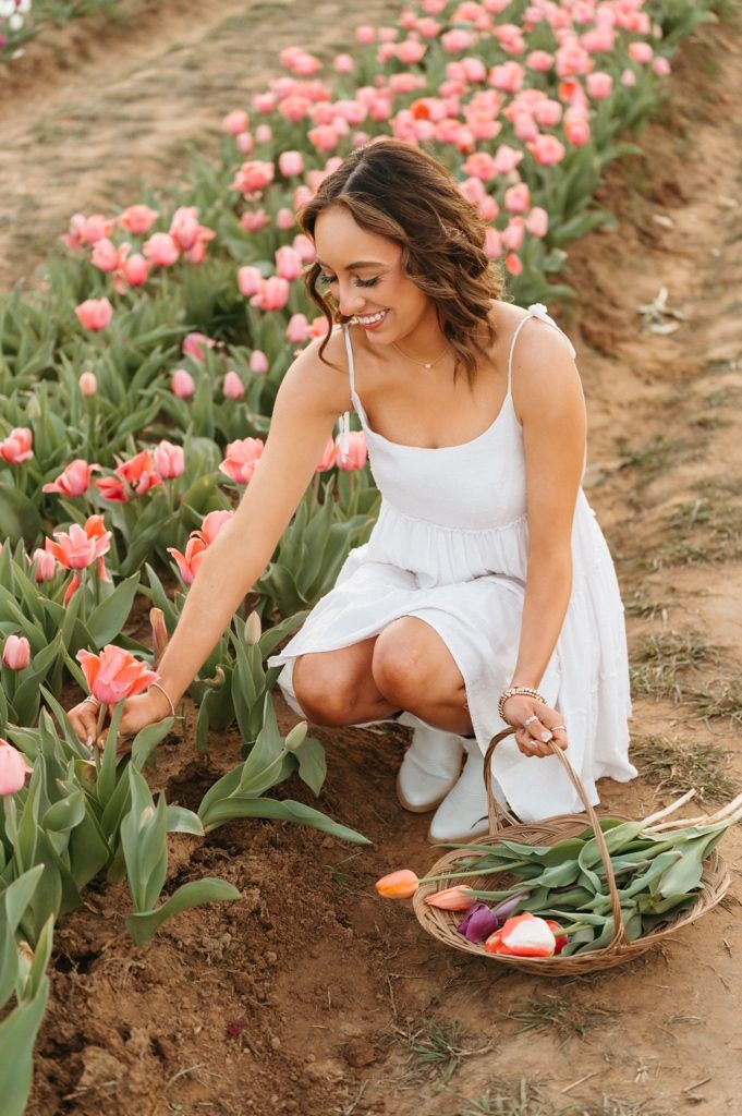 Woman in white dress kneels among pink tulips in a garden field, gathering flowers in a basket.
