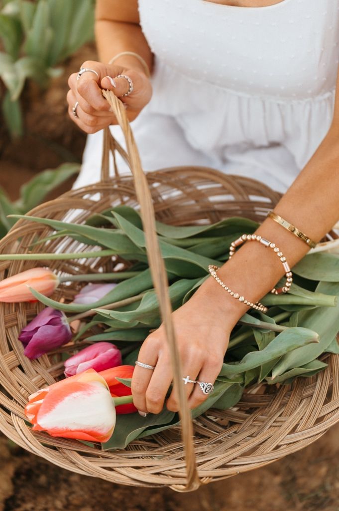 Hands with delicate bracelets arrange fresh tulips in a woven bamboo basket on garden soil.