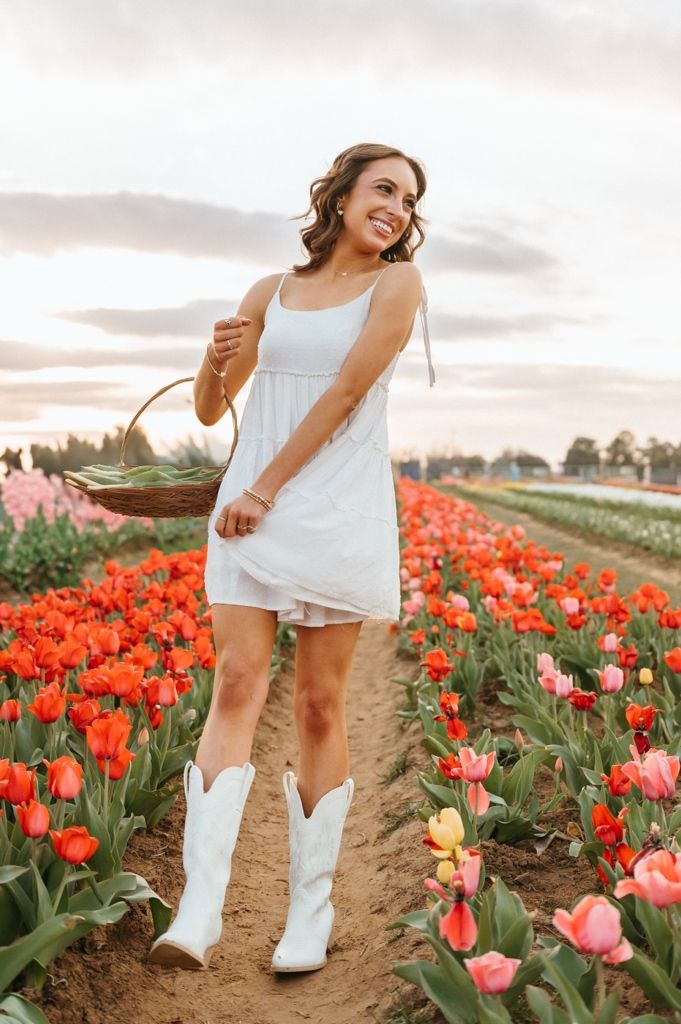 Person in white dress and cowboy boots walking through vibrant red tulip fields at sunset.