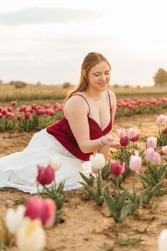 A person in a burgundy dress sits among blooming pink and white tulips in a rustic field during golden hour.