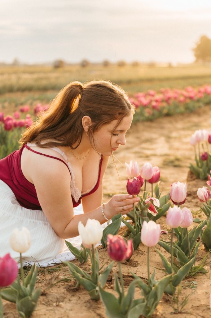 A person in a burgundy top and white skirt bends down to admire pink tulips in a field at sunset.
