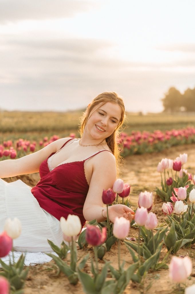 A woman in a burgundy dress reclines dreamily among pink and white tulips at sunset in a flower field.