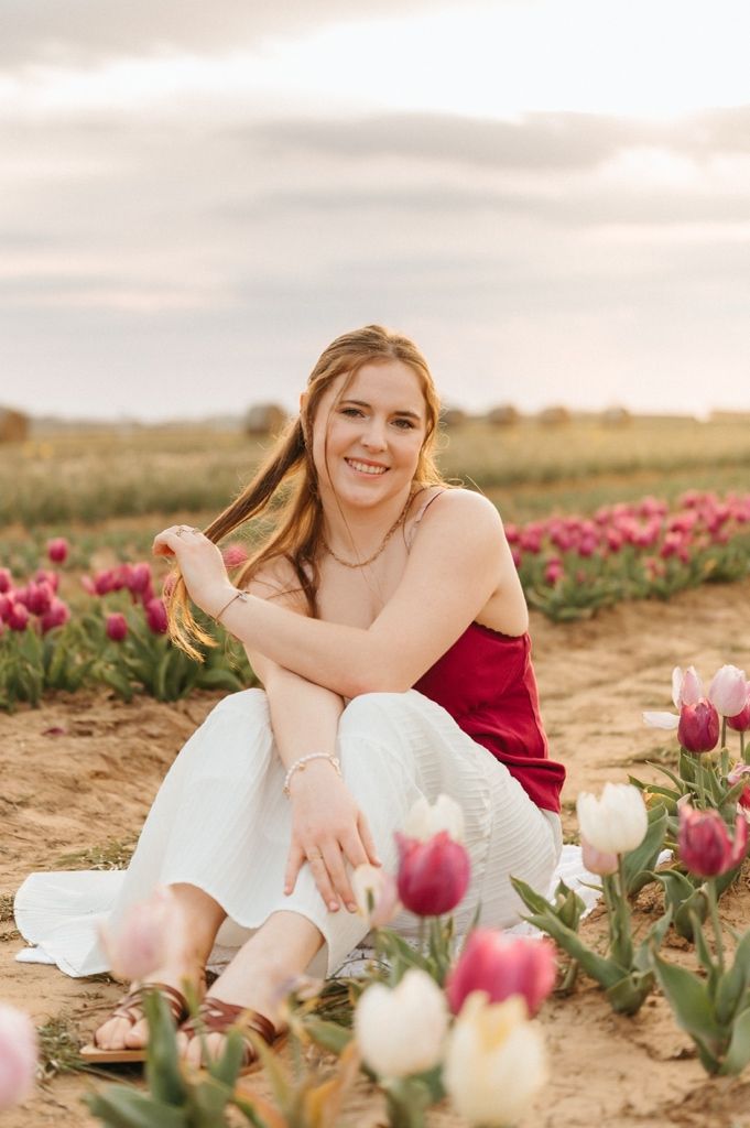 Young woman in red top and white pants sits among blooming pink and white tulips in a field at sunset.