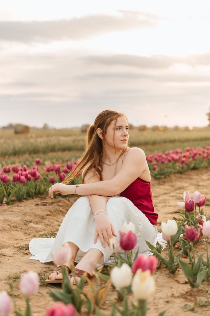 A dreamy portrait series in a tulip field at sunset shows a person in a red dress sitting among pink and white flowers.