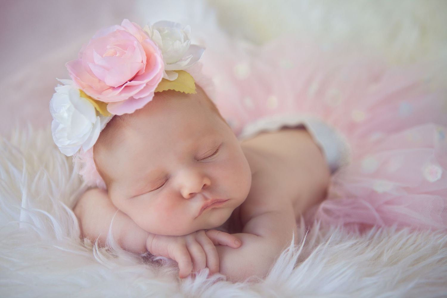 A sleeping newborn in pink tulle and lace rests peacefully on soft white fabric.