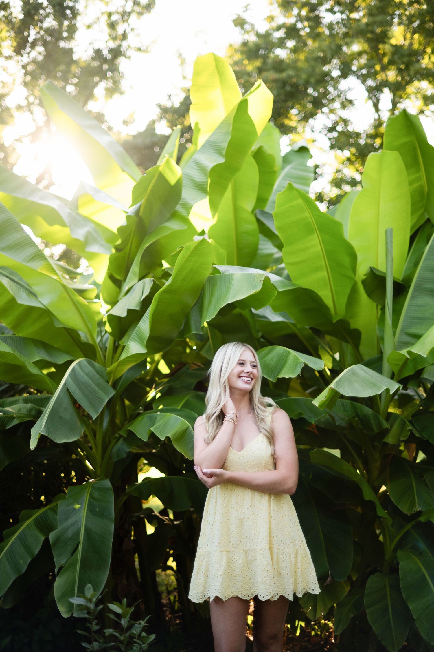 A person in a yellow sundress stands against lush green banana leaf plants in a garden setting.