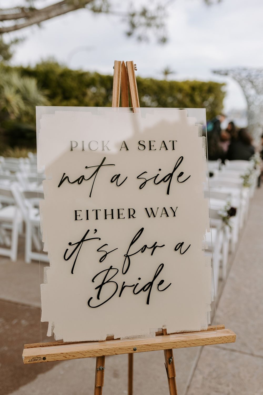 Rustic wedding ceremony sign on wooden stand reads 'Pick a seat not a side either way it's for a bride' in calligraphy.