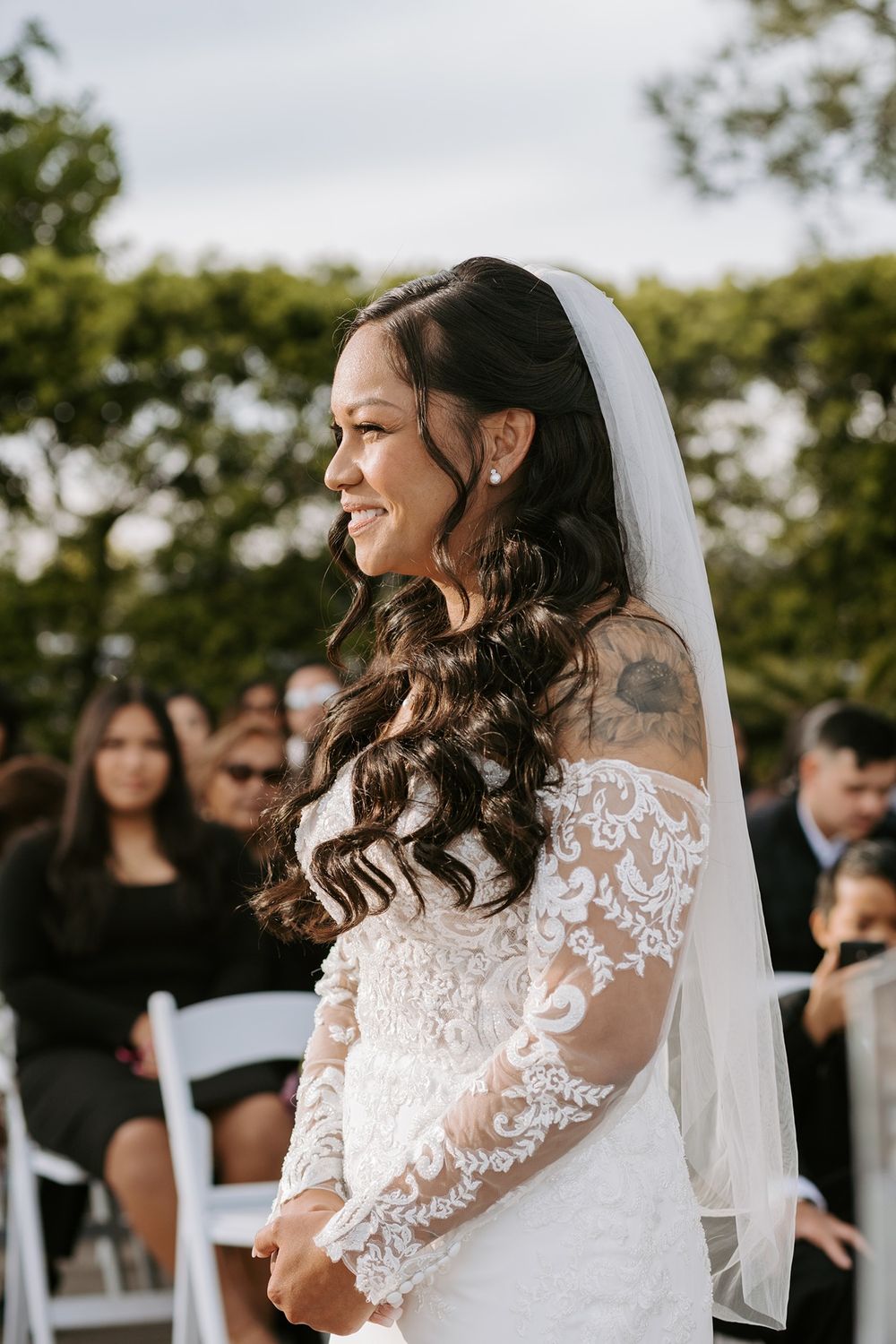 Bride in elegant lace wedding dress with long sleeves and veil smiles during outdoor ceremony at Tom Ham's Lighthouse.