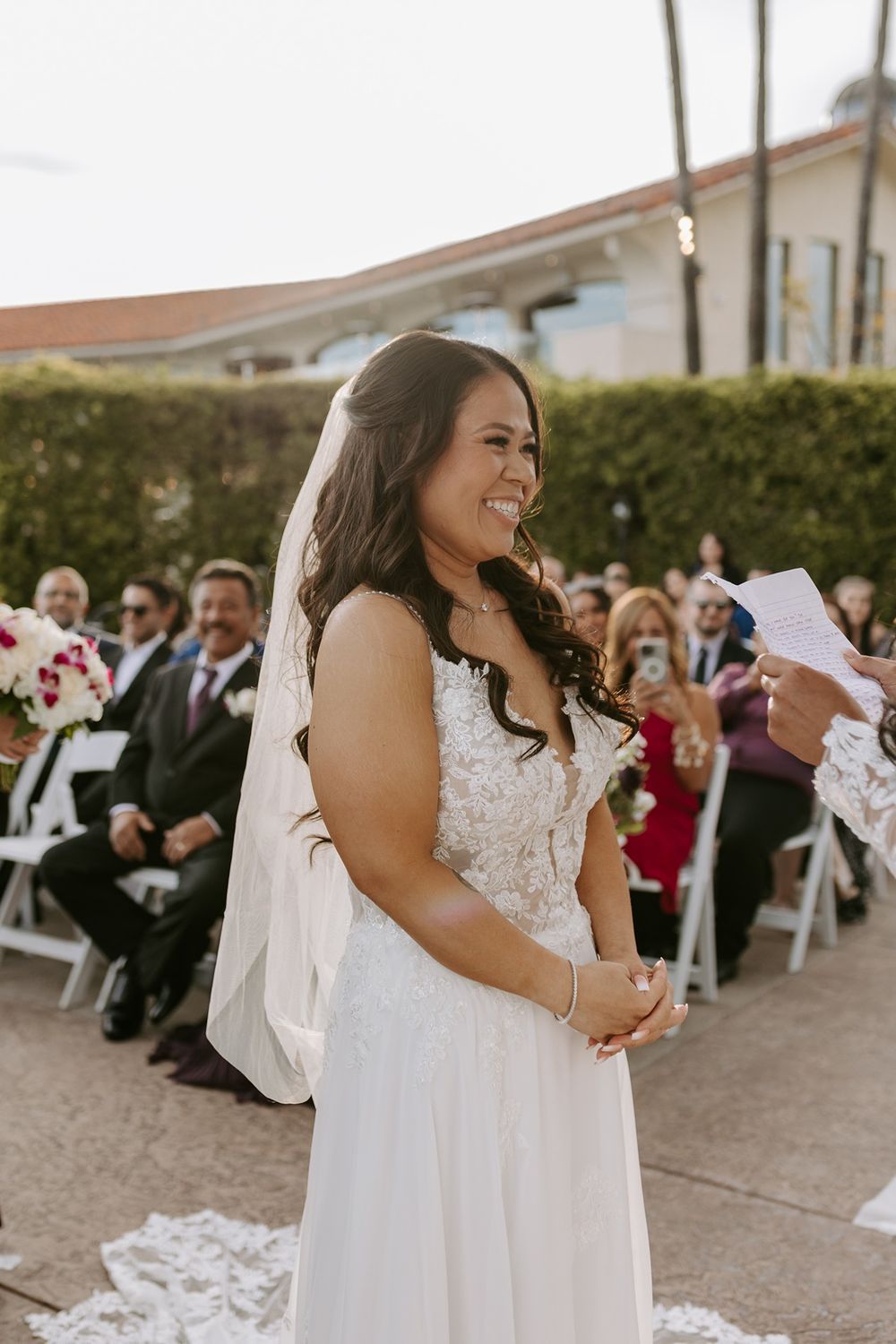 Bride in white lace dress laughs joyfully during outdoor wedding reception with guests seated in background at Tom Ham's Lighthouse.