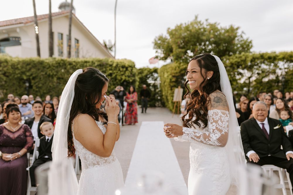 Two brides share a joyful moment during their outdoor wedding ceremony surrounded by seated guests at Tom Ham's Lighthouse.