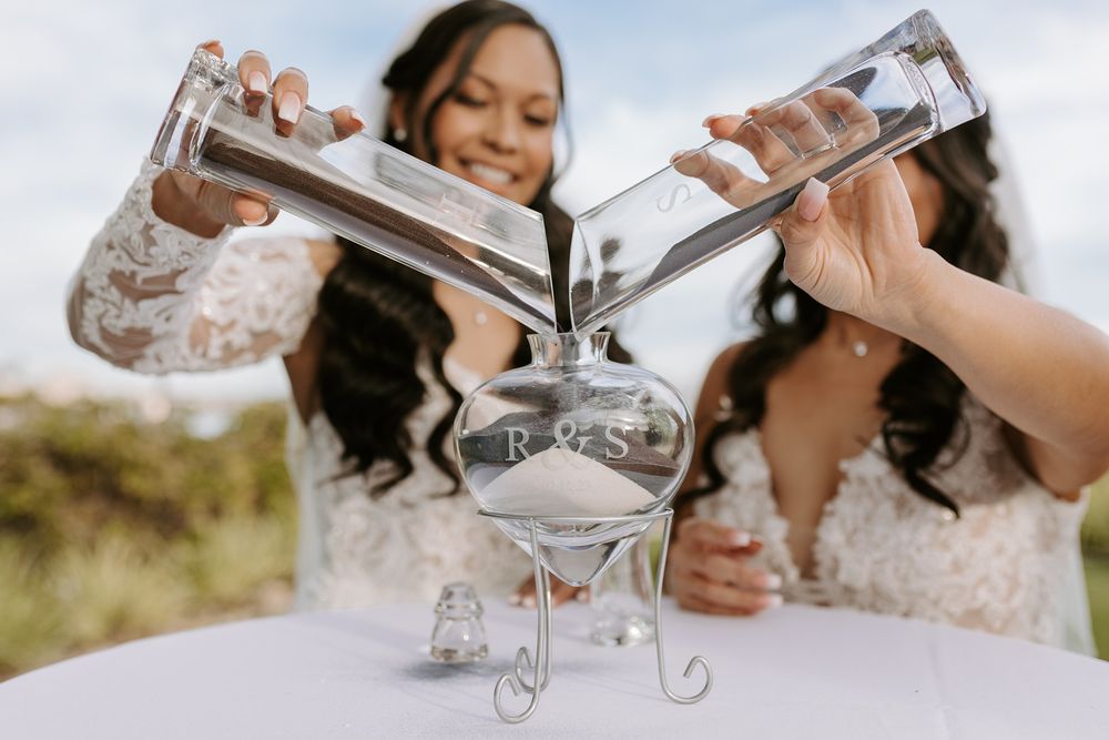 Two brides pour sand from elegant glass flutes during an outdoor wedding ceremony in natural sunlight.