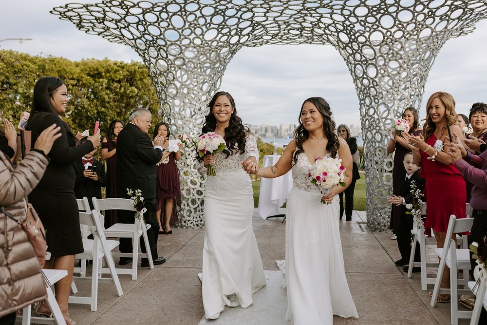 Two brides exit their wedding ceremony setup with white chairs and modern metal arch sculpture under an outdoor canopy.