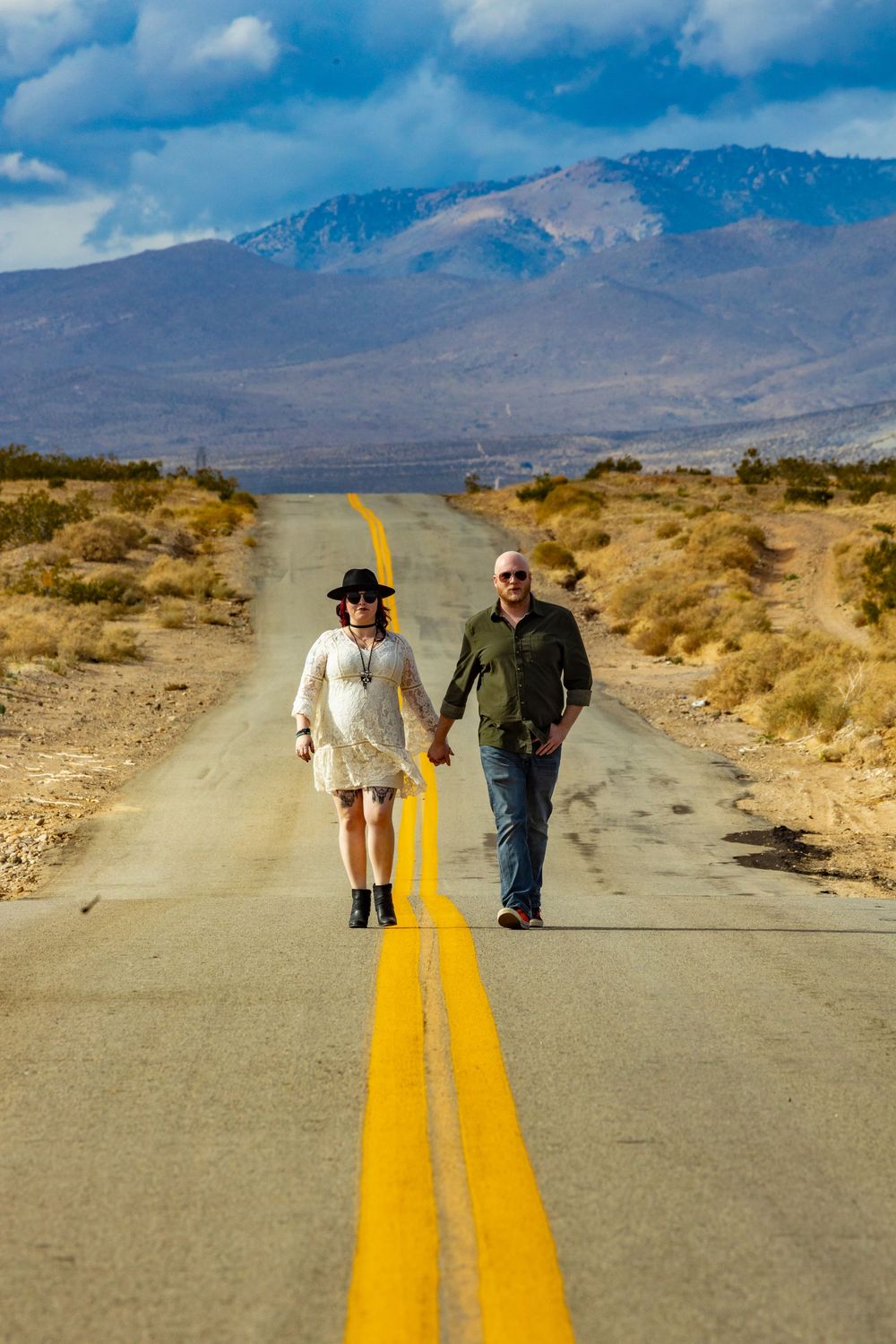 Two people walking down a desert road with mountains in the background and yellow lines painted on the pavement.