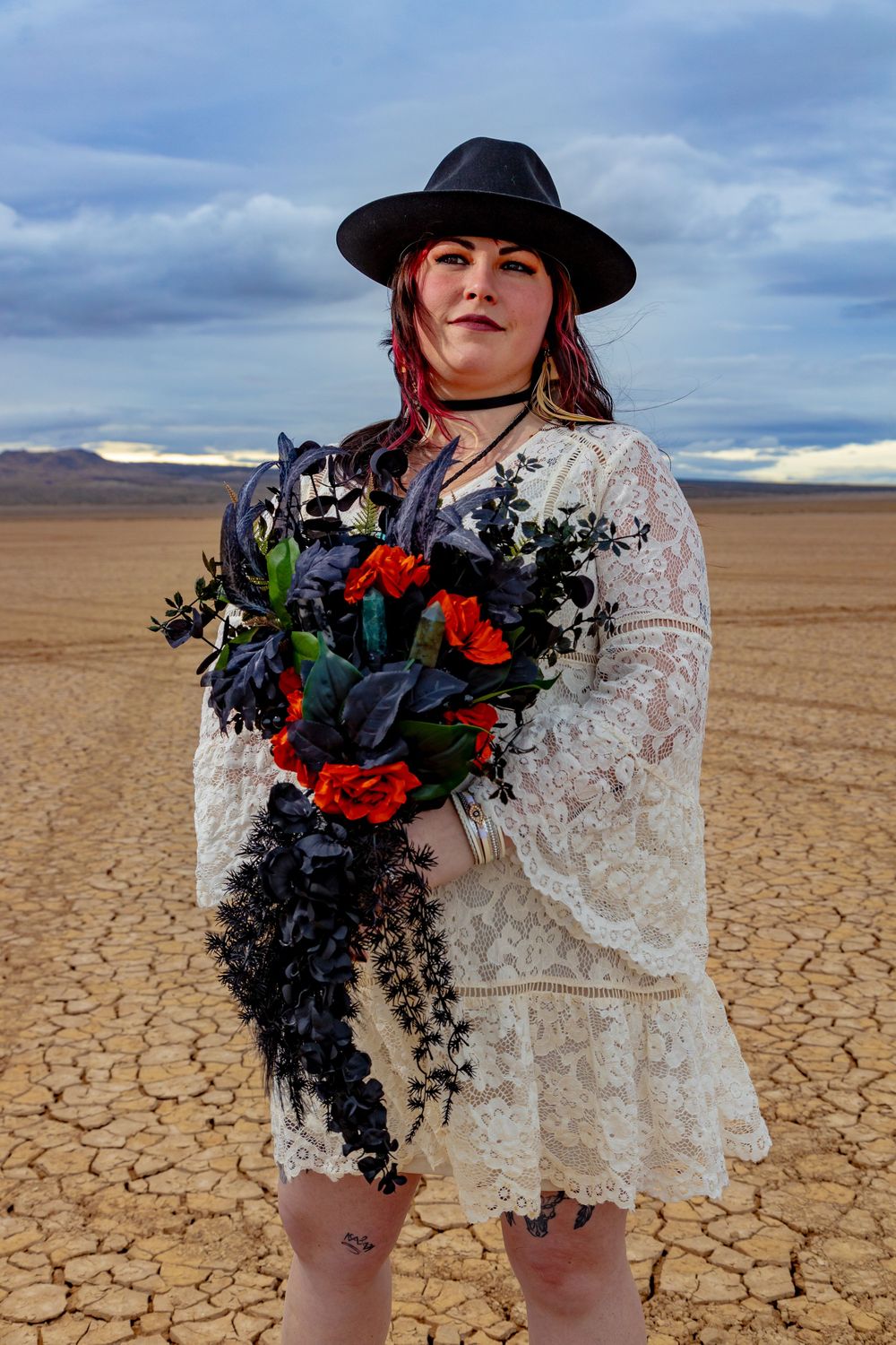 A figure in a white lace dress and black hat stands in a cracked desert landscape holding a dark floral bouquet.