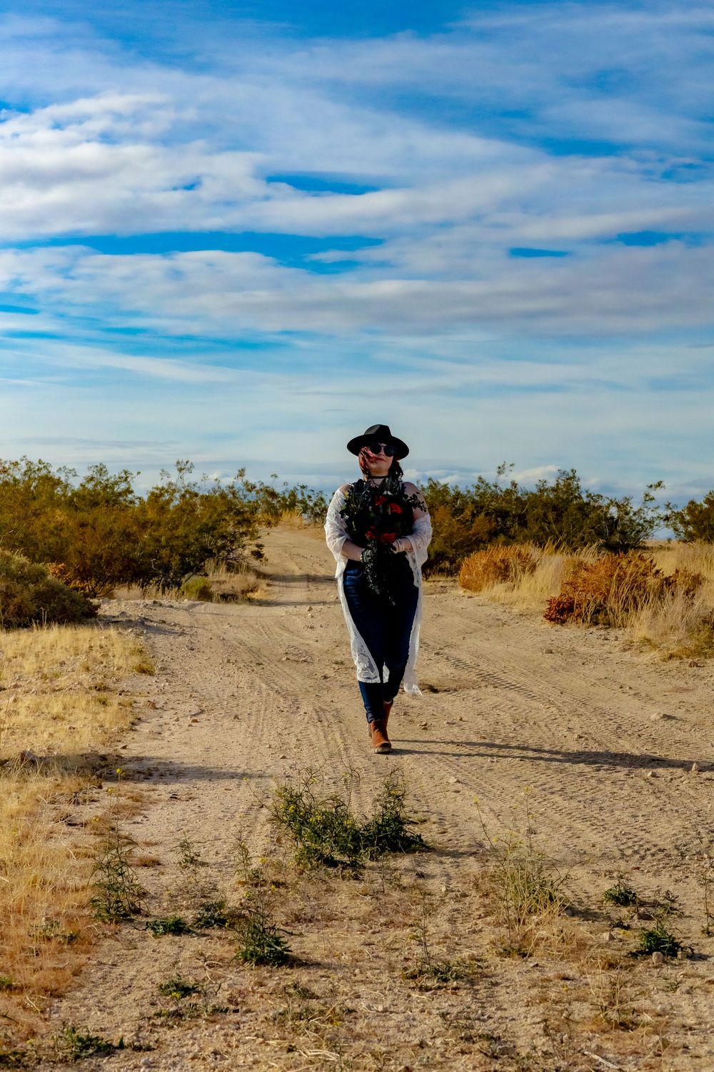 A figure in a wide-brimmed hat and long dress walks along a dusty desert trail under a bright blue sky with wispy clouds.