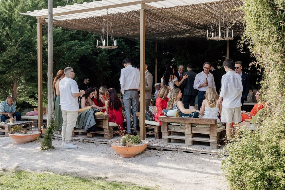 People gathered under a rustic outdoor pergola with wooden pallets and hanging lights during a casual event.