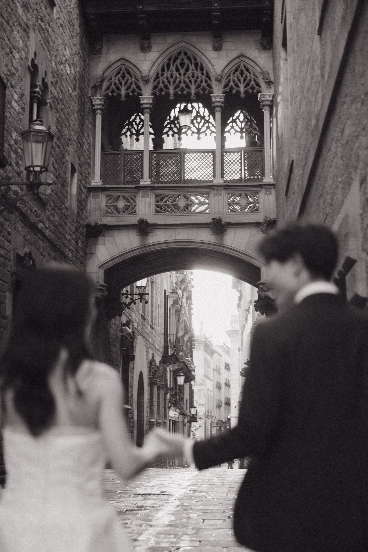 A romantic black and white photograph shows silhouettes of people walking under an ornate Gothic archway in Venice.