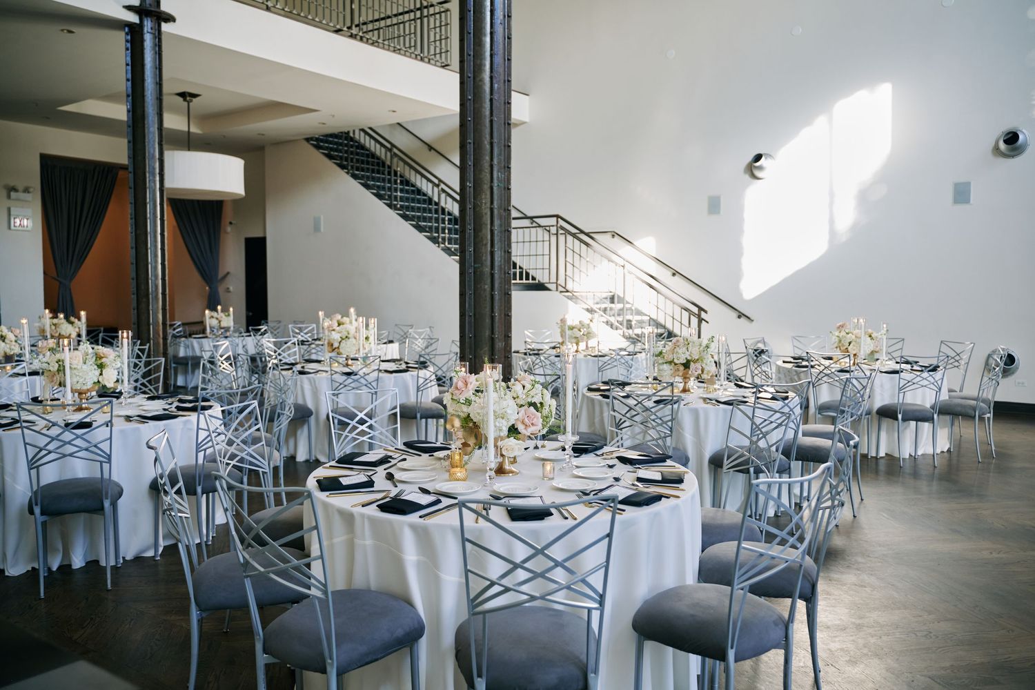 Modern wedding reception venue with elegant white and gray table settings arranged in rows under industrial-style ceiling.