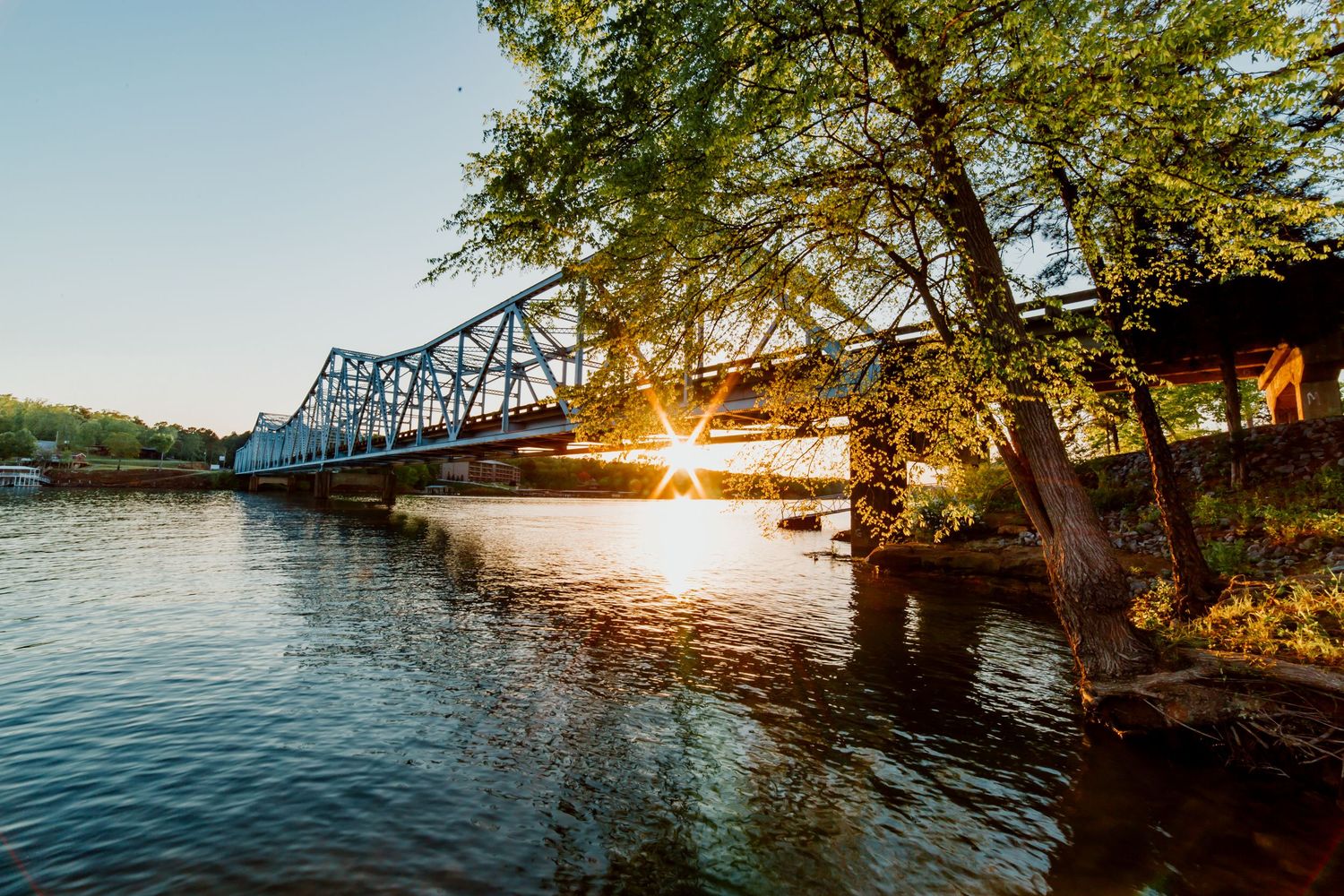 Panoramic view of a railroad bridge over calm water at sunset with golden sunlight filtering through trees.
