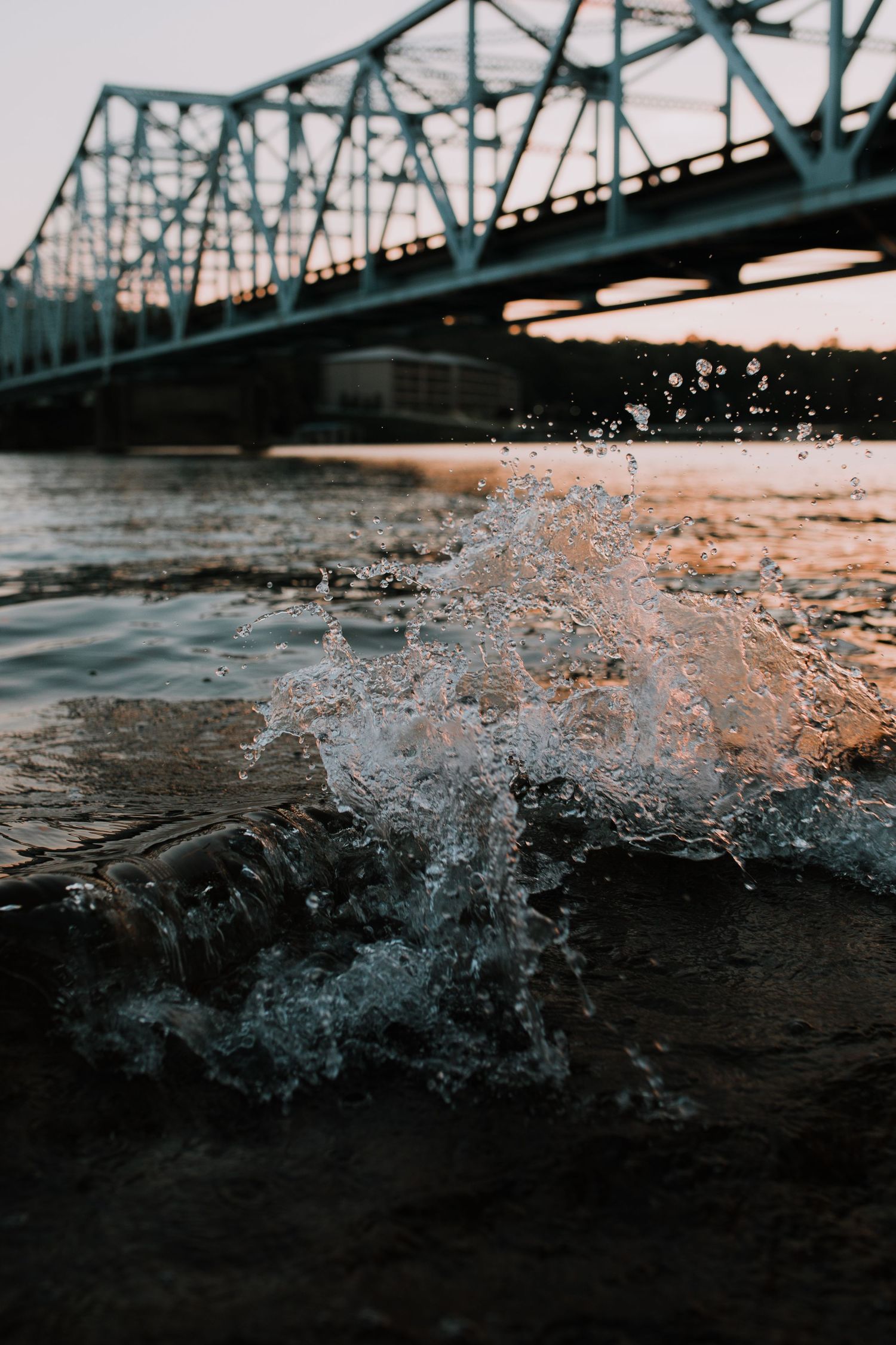 Water splashes against dark sand at sunset with a bridge silhouetted in the background.